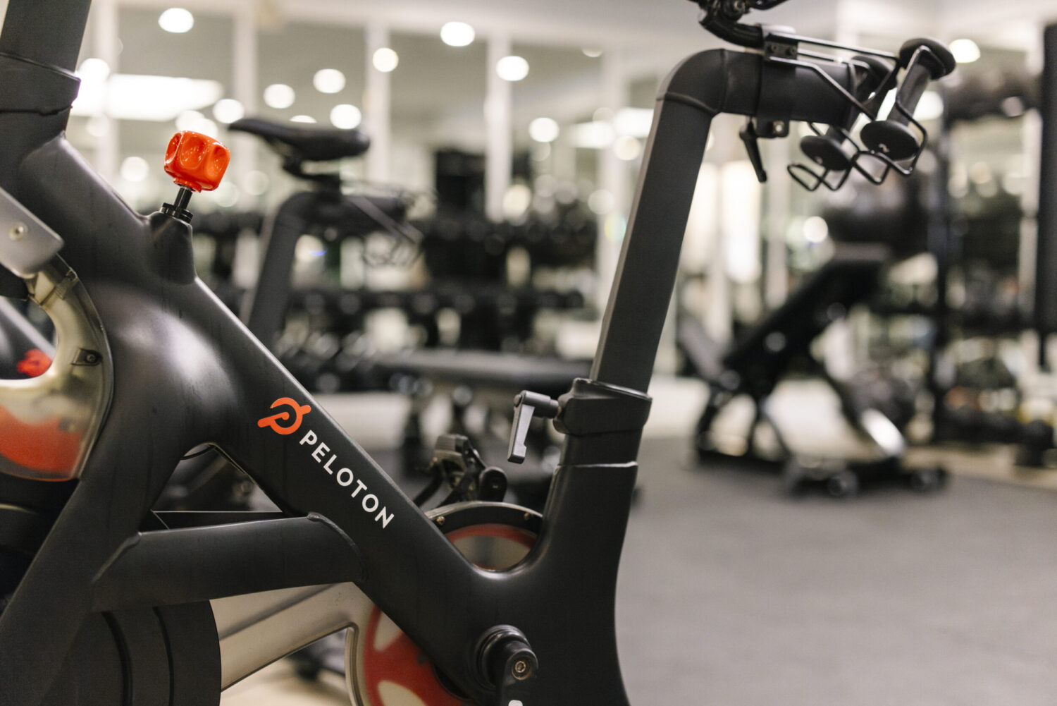 Close-up of a Peloton exercise bike in a gym, with exercise equipment visible in the background.