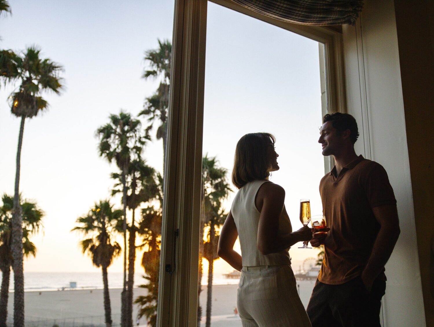 A couple by a window holding drinks, enjoying a sunset view of palm trees and Santa Monica beach.