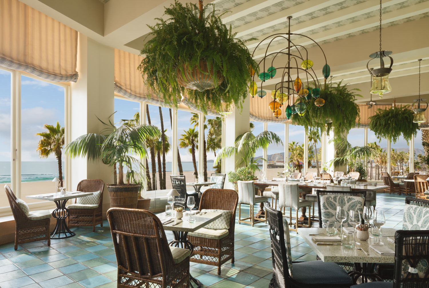Bright luxury restaurant with wicker chairs, large windows, and hanging plants overlooking a beach.