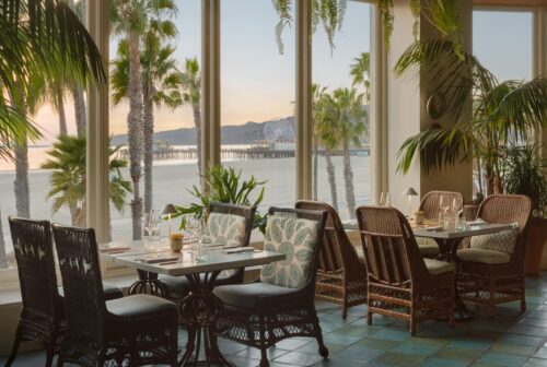 Dining area with wicker chairs and tables set by large windows overlooking Santa Monica beach.