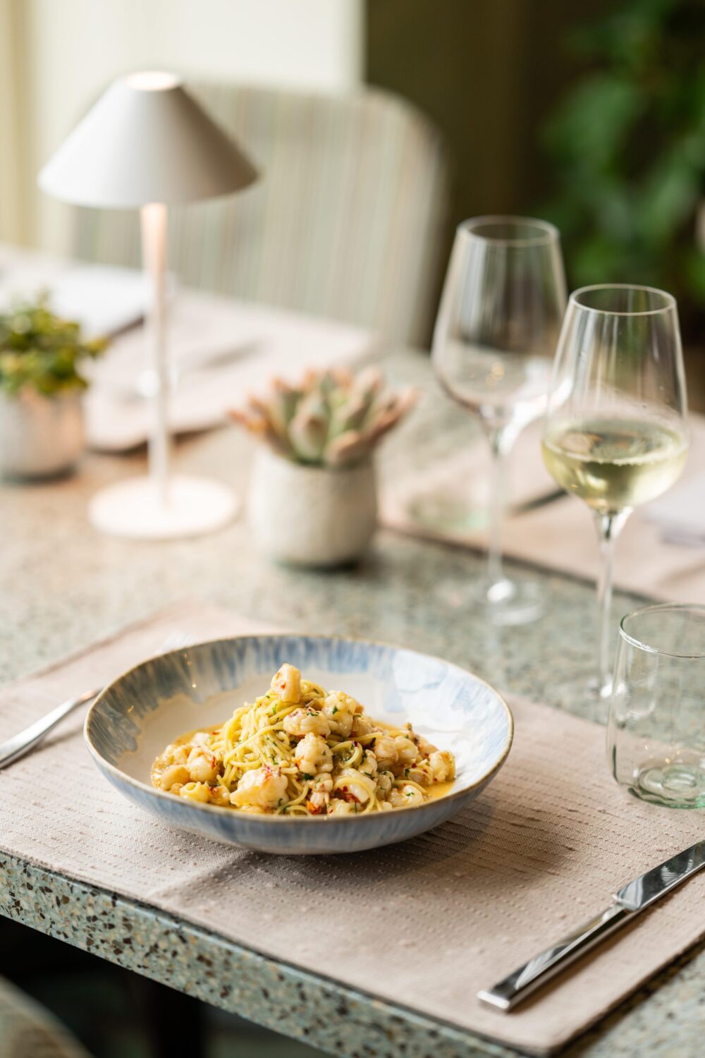 Bowl of pasta with shrimp on a speckled countertop, accompanied by glasses of white wine.