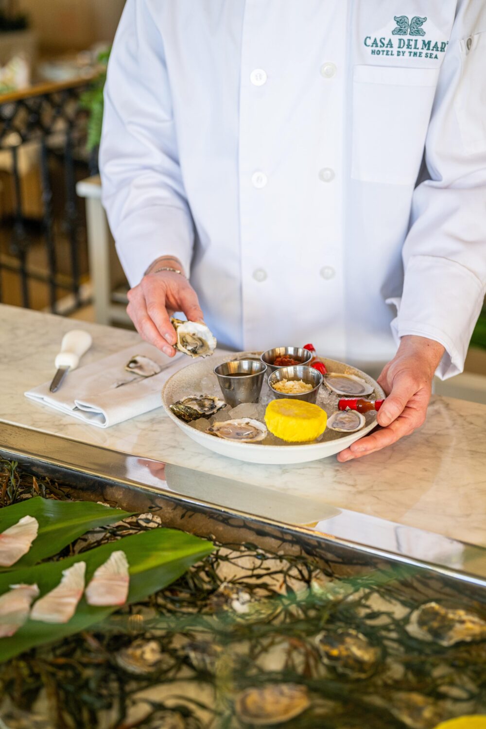 Chef in a white uniform holding a plate of oysters with various condiments, standing at a counter.