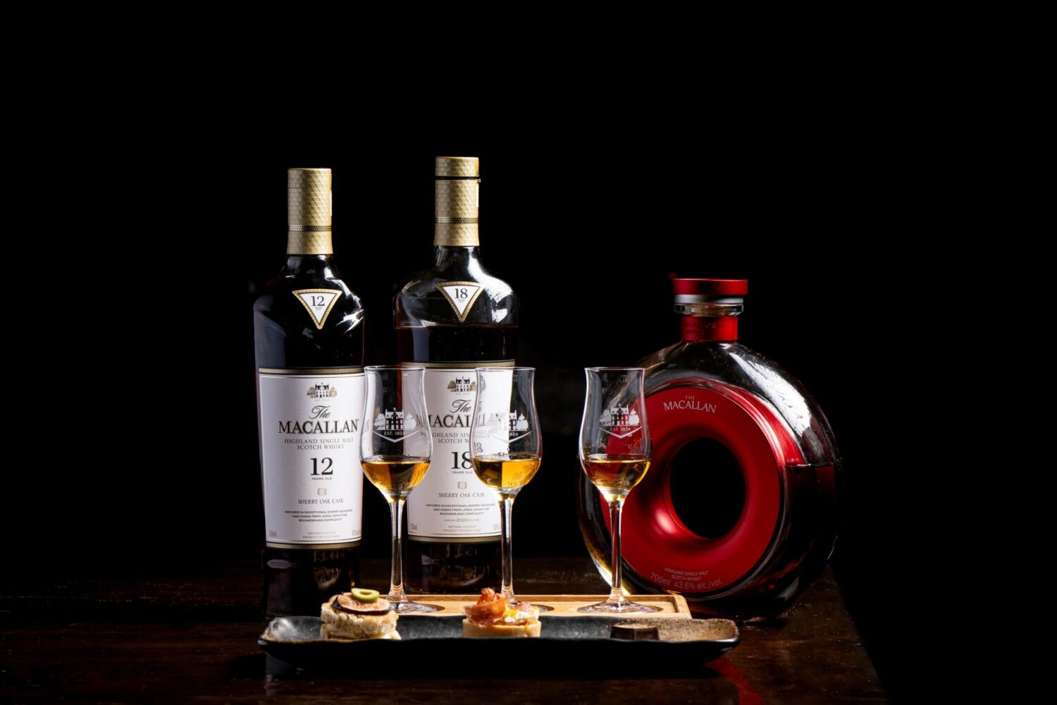 Two tall bottles of Macallan whisky, a round red decanter, & three whisky glasses on a wooden table.
