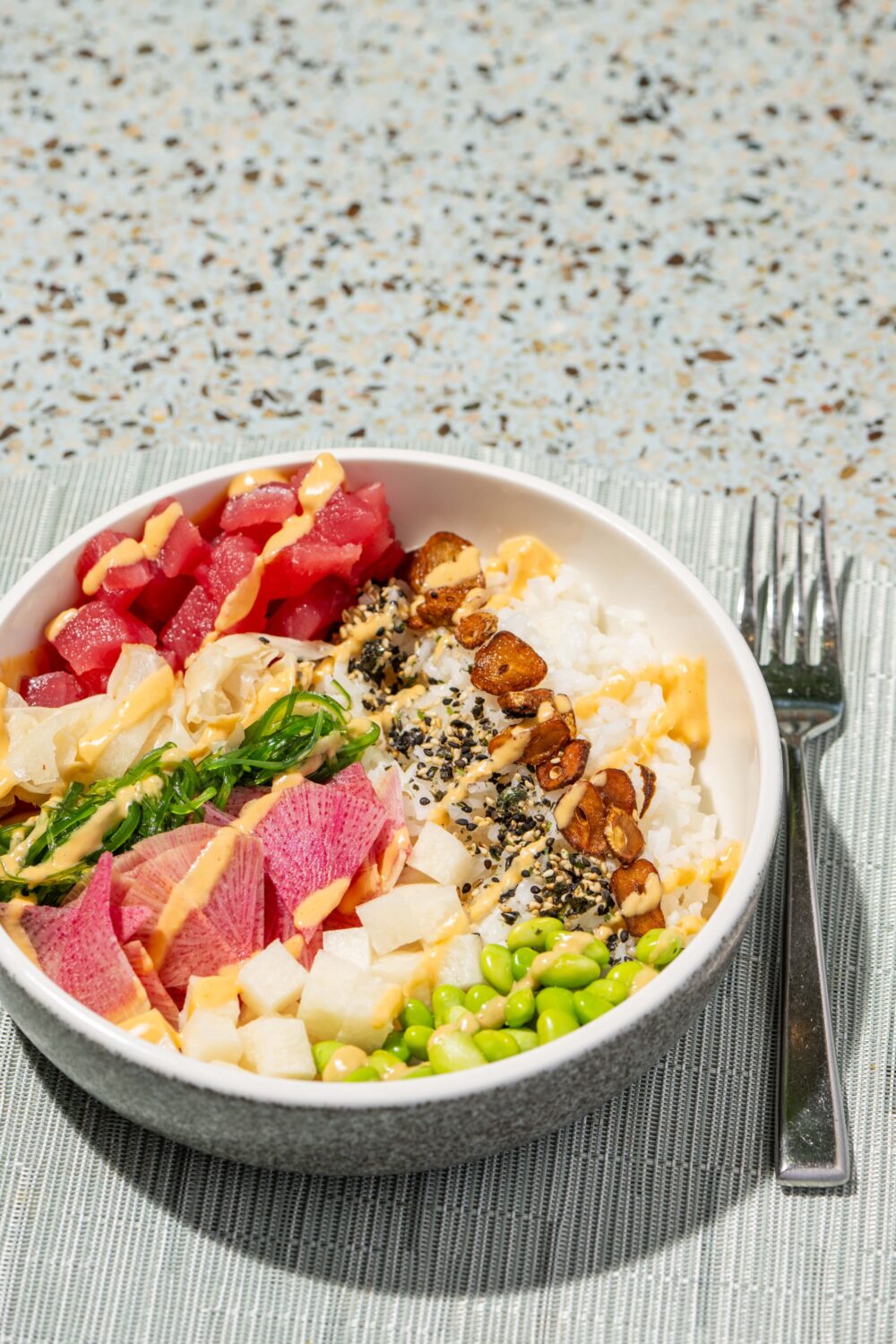 Bowl of poke with tuna, edamame, rice, seaweed, nuts, and various toppings, accompanied by a fork.