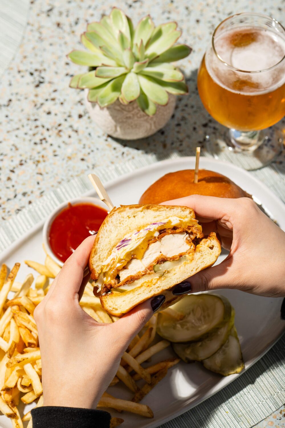 Hands holding a chicken sandwich with fries, pickles, and a glass of beer on a speckled table.