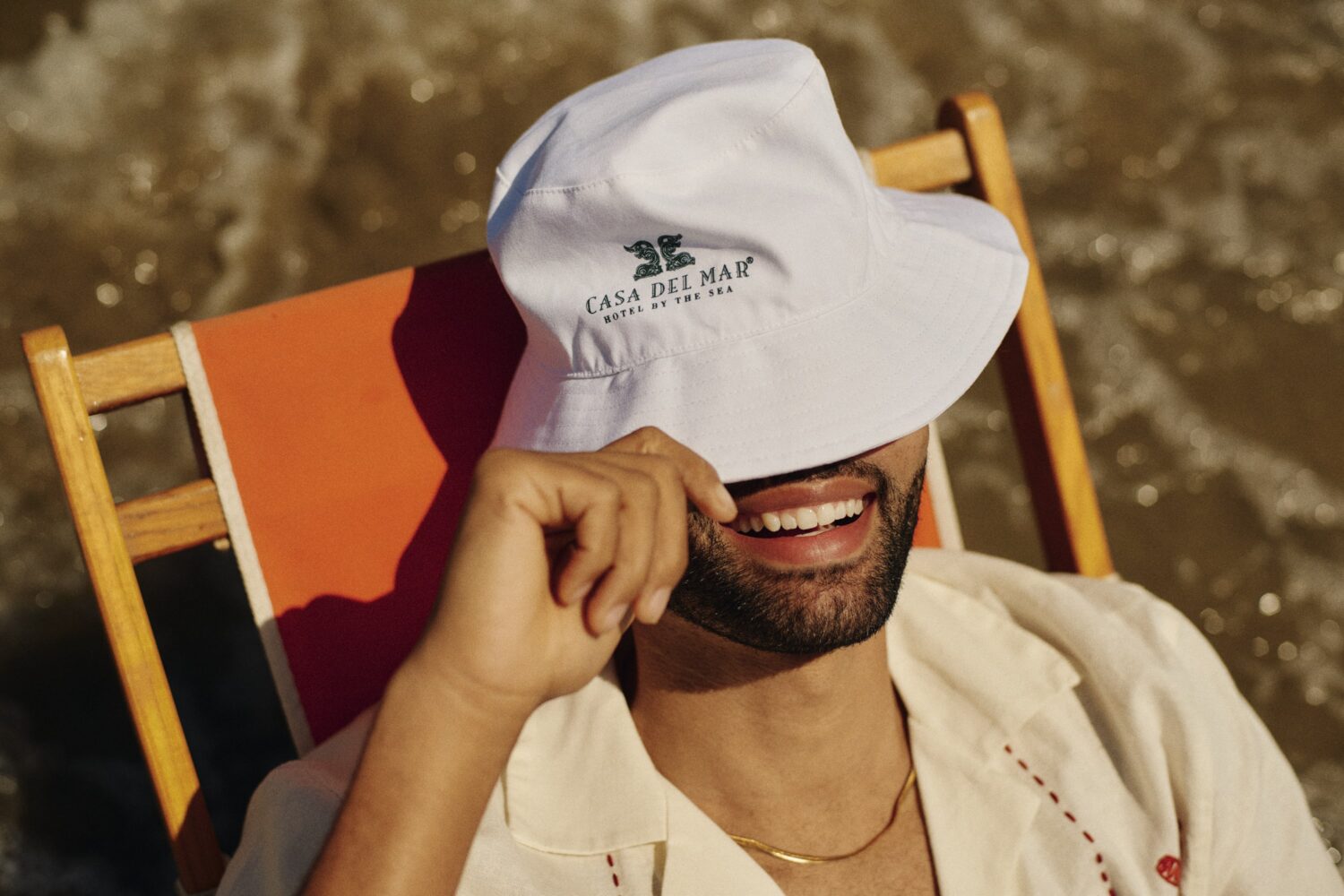 Man on the beach, smiling with a Casa del Mar bucket hat over his eyes.