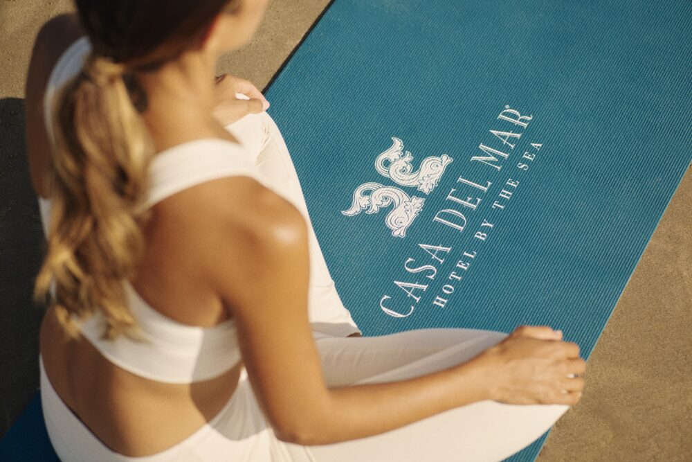 Casa del Mars yoga mat on the beach, with a woman sitting cross-legged on the mat