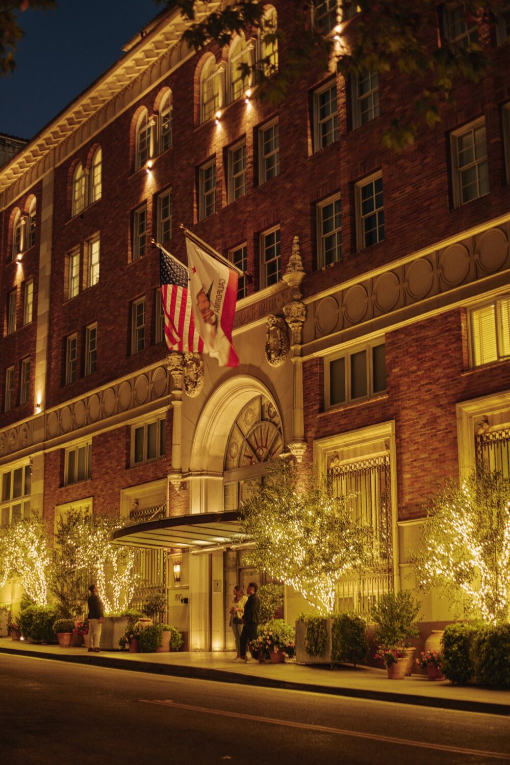 Nighttime exterior of Casa del Mar Hotel, with lit trees, flags, and a couple at the door.