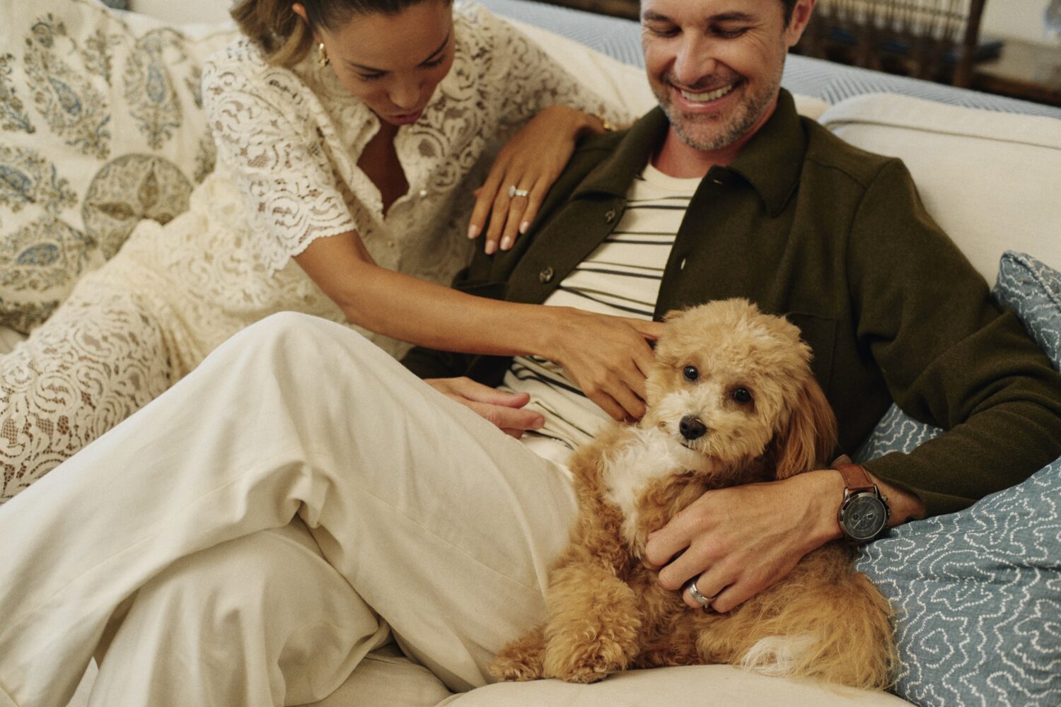 a man and a woman sitting on a couch, smiling and petting a cute tan and white goldendoodle puppy