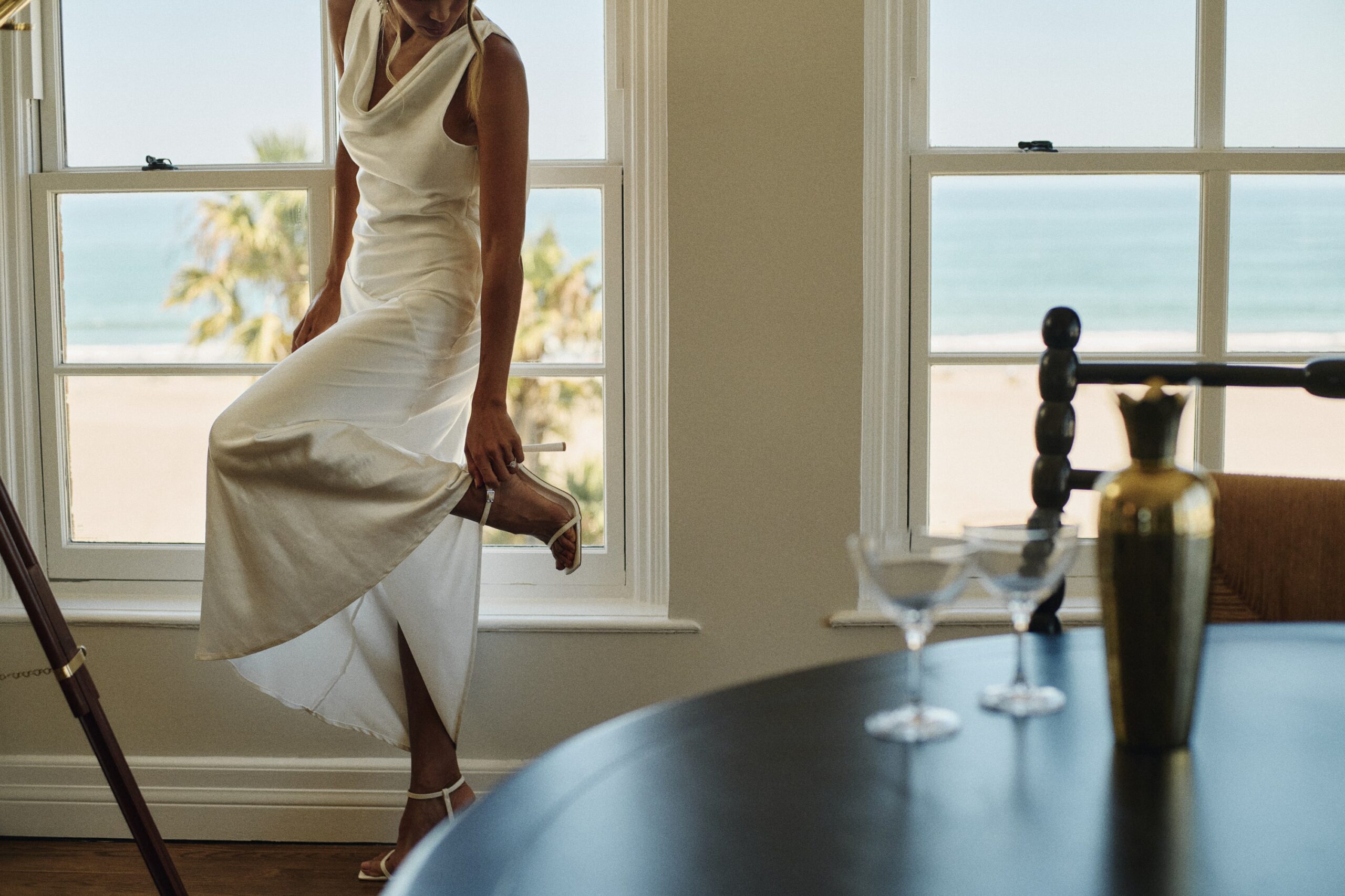 woman in her wedding dress holding her heel in one hand, posing by the window with an ocean view