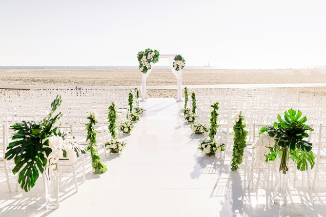 Elegant wedding setup at Casa Del Mar Hotel with a white floral arch, plants & rows of white chairs.