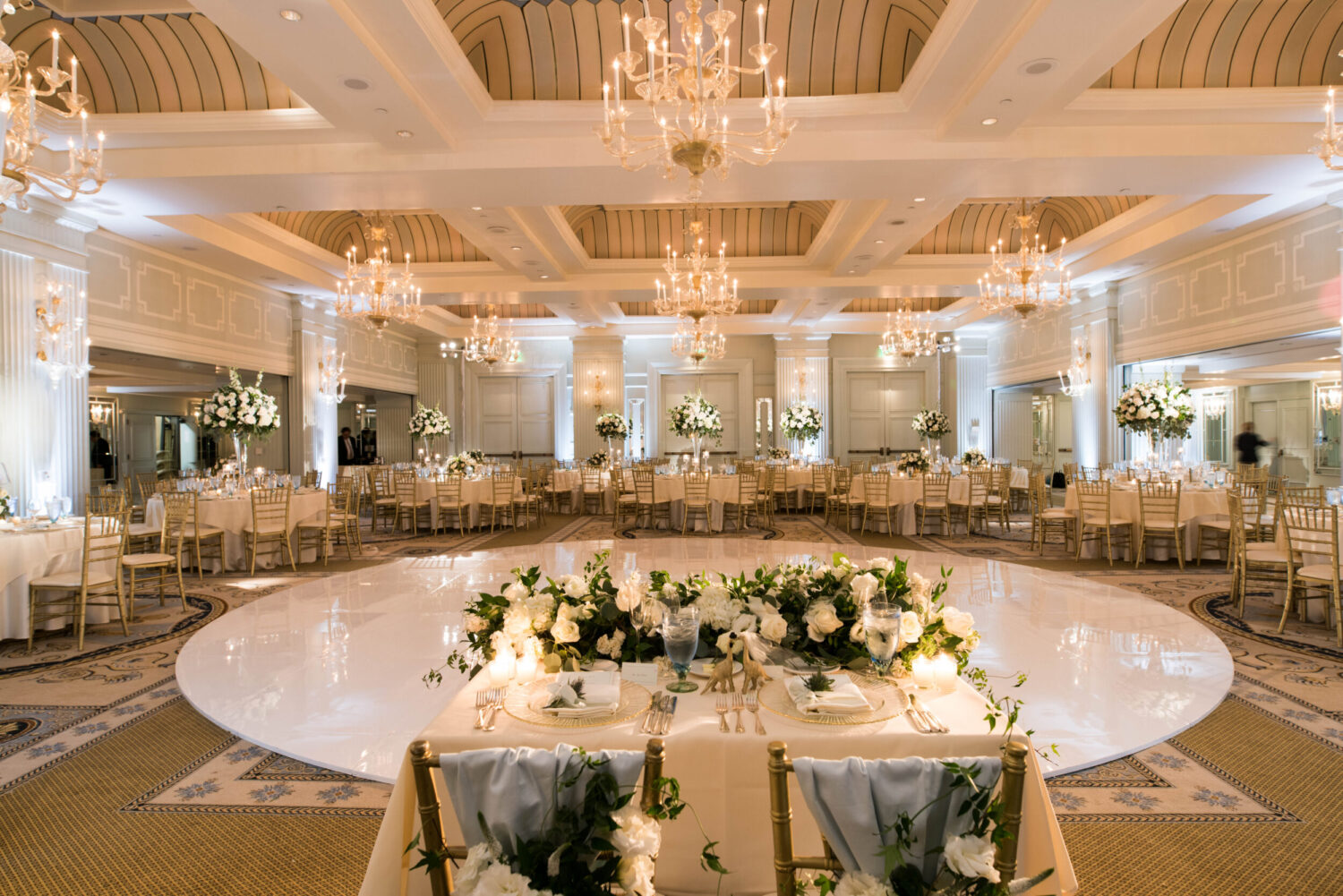 Wedding reception featuring a ball room with chandeliers, a white dance floor and decorated tables.