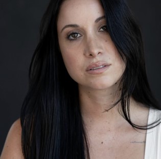 A woman with dark hair in a white top looks into the camera against a dark background.