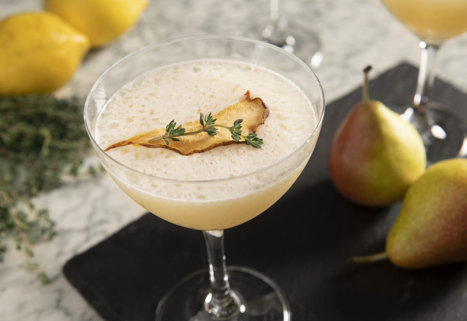 Foamy cocktail in a coupe glass, garnished with pear slice and thyme, surrounded by lemons.