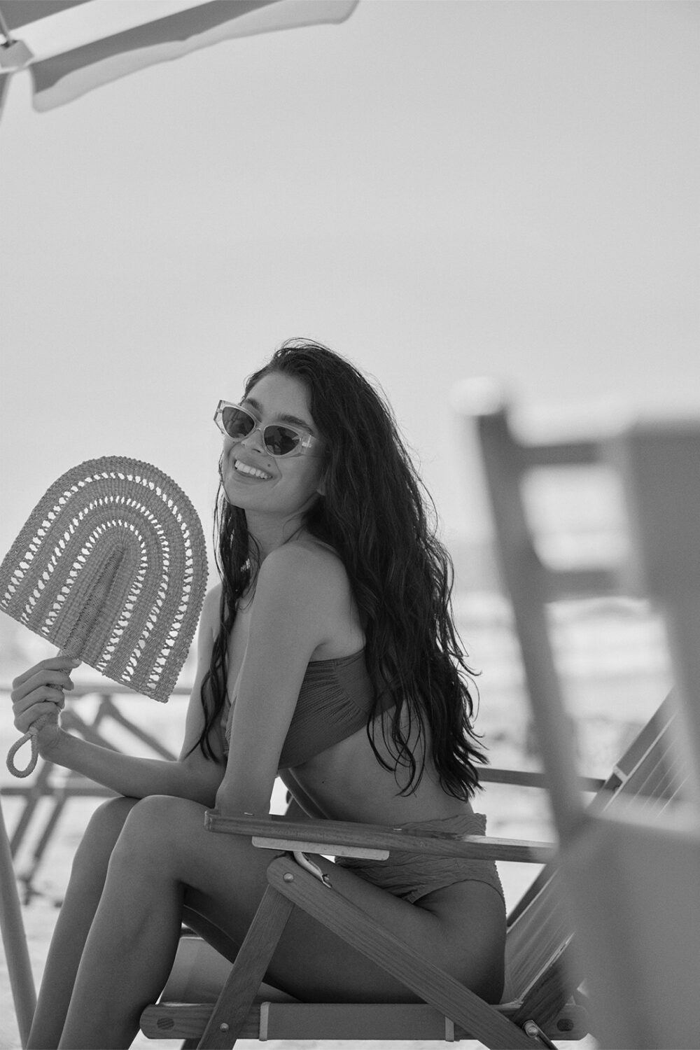 b&w image woman on the beach, sitting in a beach chair fanning herself with a crochet fan