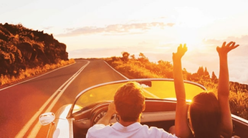 A couple driving a convertible at sunset, arms raised, enjoying the open road.