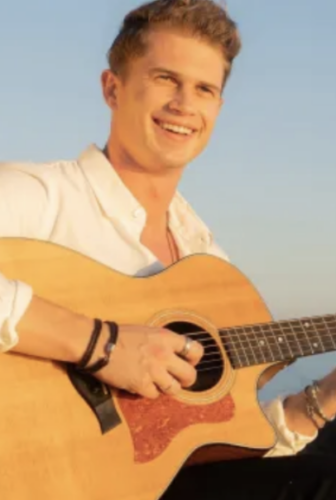 A man smiling while playing an acoustic guitar outdoors under a clear blue sky.