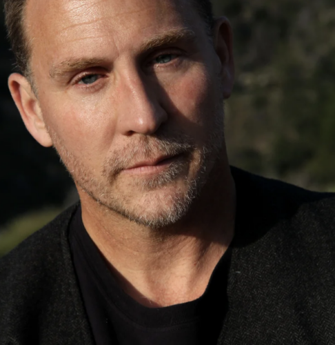 A man with short hair and a beard, wearing a black shirt, looking thoughtfully into the distance.