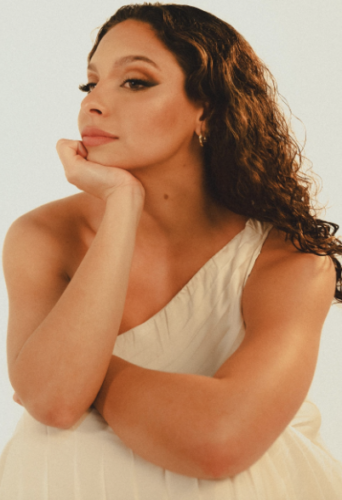 A woman with curly hair in a white dress, resting her chin on her hand and looking thoughtfully.